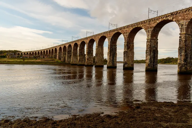 The tall stone arches of the Royal Border Bridge striding across the River Tweed at low tide, with overhead line masts along the top