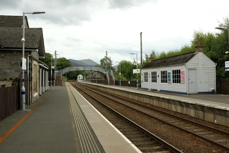 Blair Atholl station's two platforms with a white timber waiting room, a lattice footbridge and hills beyond