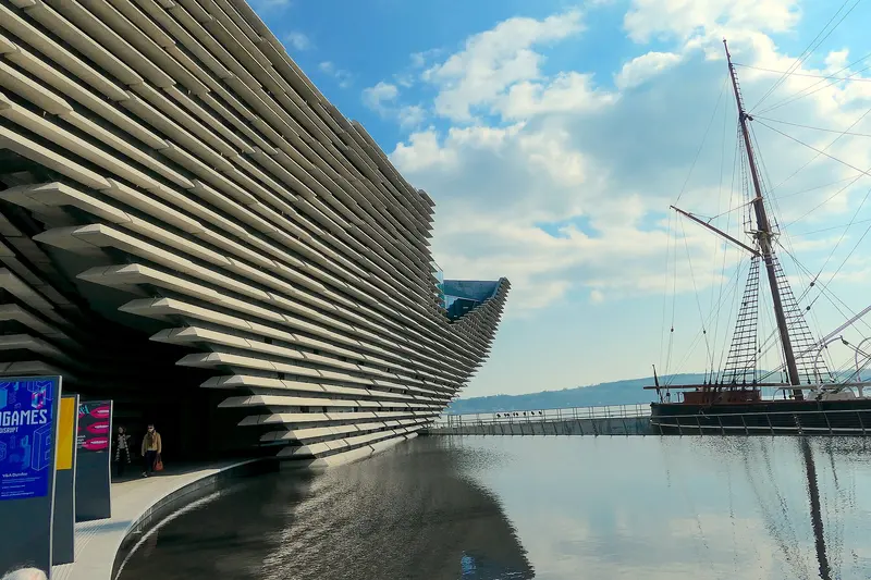 The layered concrete facade of V&A Dundee reflected in a shallow pool, with the masts of RRS Discovery alongside