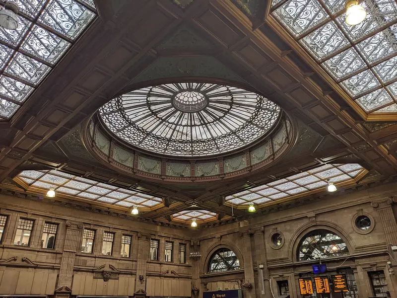 The ornate glazed dome and coffered ceiling above Waverley's booking hall, with carved stone walls and a departure screen below