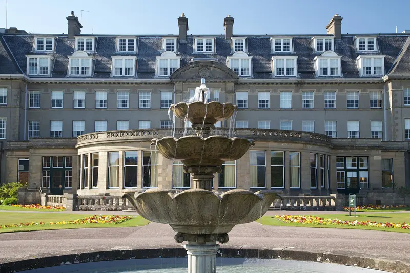 The grand stone front of the Gleneagles hotel with a tiered fountain and flower beds in front