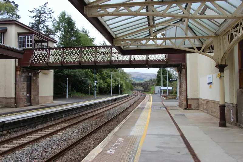 Looking along a platform at Gleneagles under an ornate glazed canopy, with a lattice footbridge across the tracks
