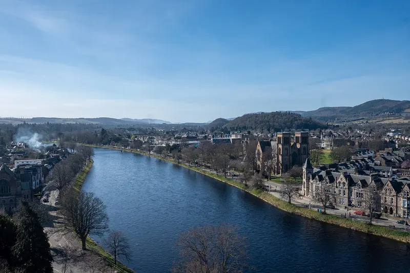 The River Ness curving through Inverness, lined with trees, churches and stone houses, with hills beyond