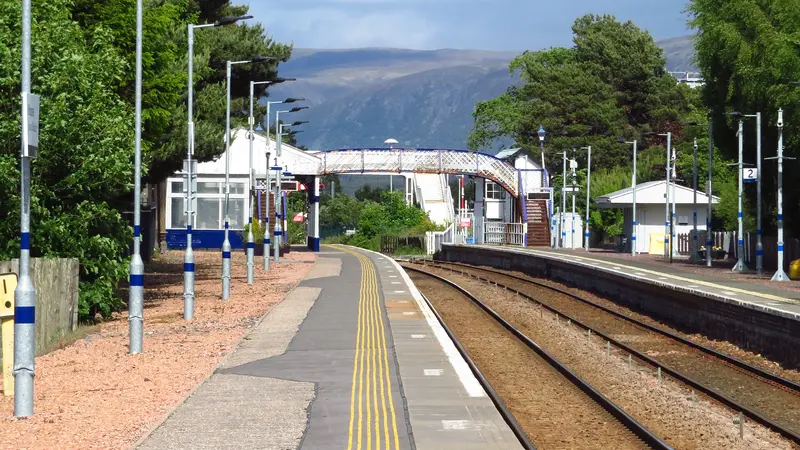 Kingussie station's two platforms and lattice footbridge, with pine trees and a broad hill behind