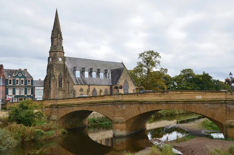 A three-arched stone bridge over the River Wansbeck in Morpeth with a church spire behind