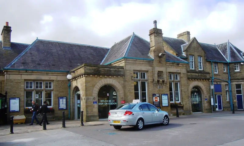 The stone front of Skipton station on Broughton Road, with gabled wings, tall chimneys and a taxi waiting outside